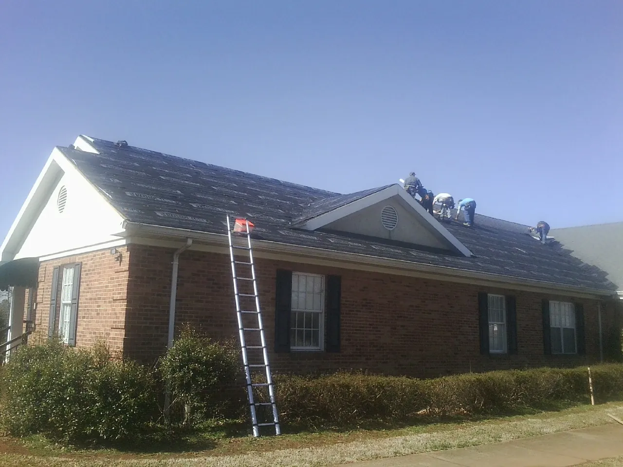 Crew tearing shingles off a brick ranch roof with a ladder set — Metal Roof Repair work by our crew in Schenevus