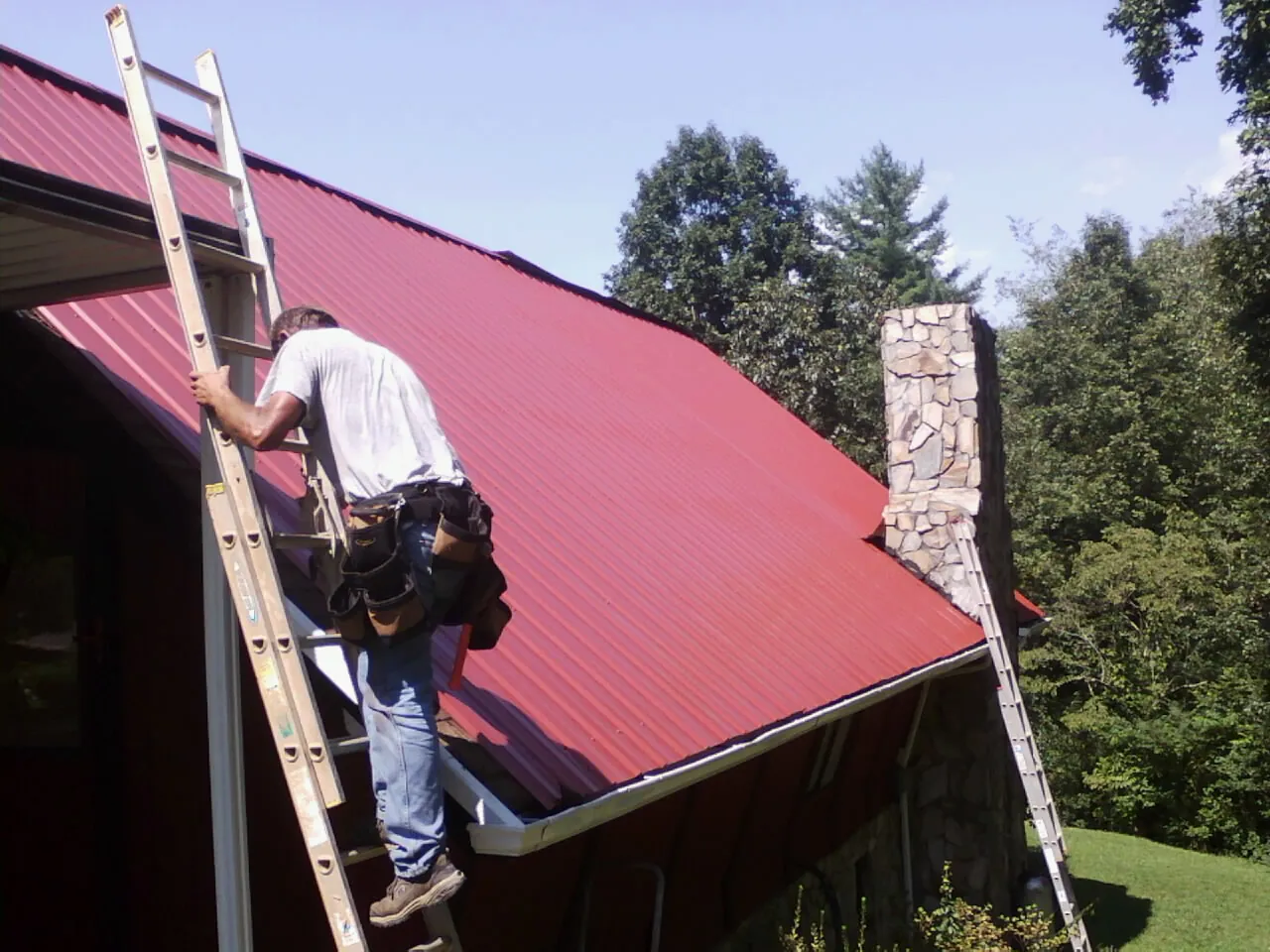 Roofer on a ladder beside a red metal roof and stone chimney — Soffit & Fascia Repair by Roofing Brothers in Schenevus